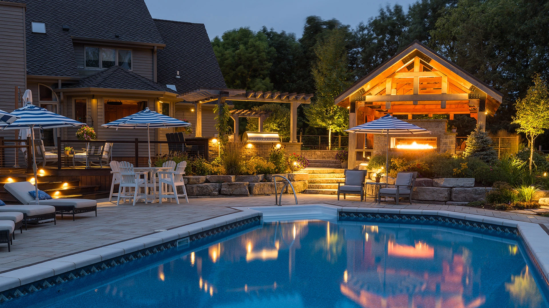 Night view of a landscaped backyard with a swimming pool, lounge chairs, and a pavilion with a fireplace in Wauwatosa, WI.