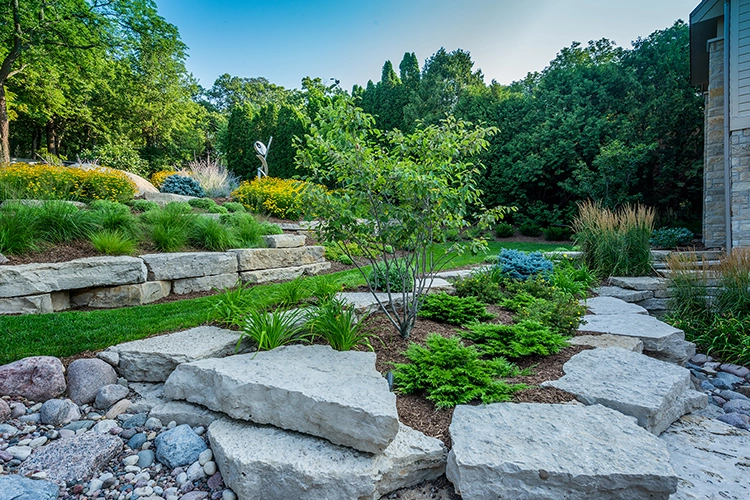 Beautiful garden with stacked stone walls, colorful flowers, and greenery, featuring a modern sculpture in Whitefish Bay, WI.